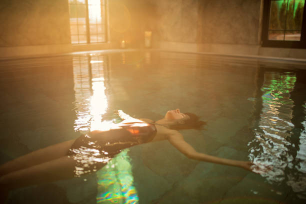 Young pregnant woman relaxing and enjoying on health spa poolside. She is floating on water in swimming pool, wears one piece swimsuit and looks beautiful, serene and carefree. Sunlight from background make beautiful colors with heat haze inside.