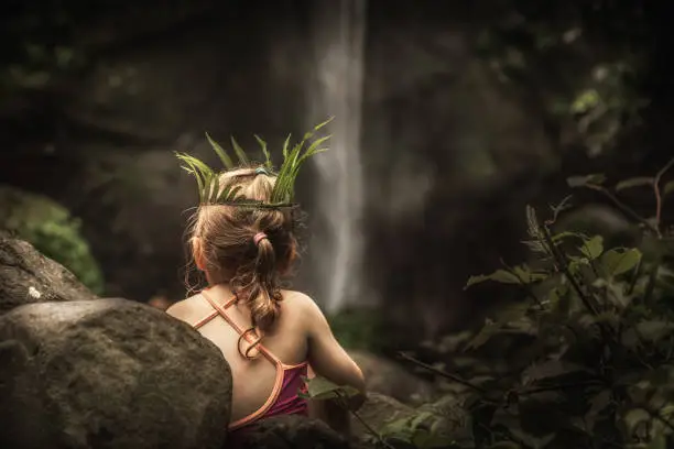 Photo of Lonely pensive child girl in wild forest looking at waterfall with plant wreath on her head as exploring wild nature concept