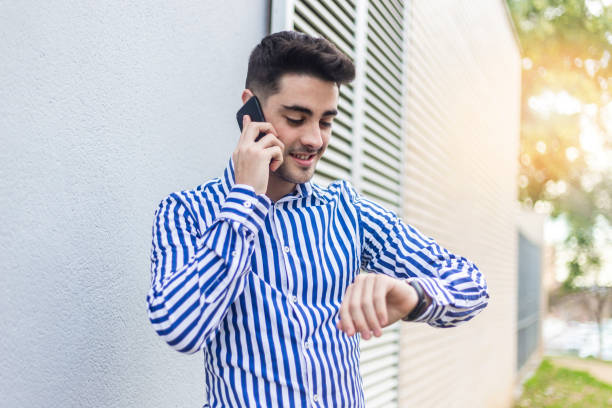 istock Young man standing outdoors while looking his wrist watch and talking by phone 1205848689