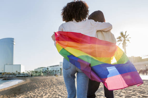 couple of gay girls walking with the rainbow flag interracial couple of gay girls walking in front of the sea at Barcelona wrapped in the rainbow flag, symbol of the struggle for homosexual rights, concept of sexual freedom and racial diversity lesbian pride stock pictures, royalty-free photos & images