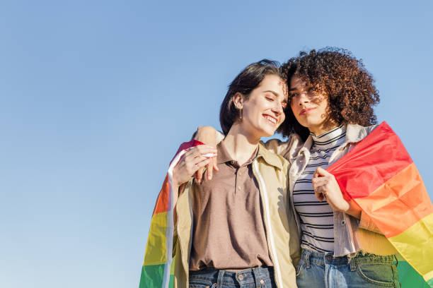 gay couple of women embracing with a rainbow flag interracial gay couple of lovely women embracing wrapped in the rainbow flag, symbol of the struggle for homosexual rights, concept of sexual freedom and racial diversity lesbian pride stock pictures, royalty-free photos & images
