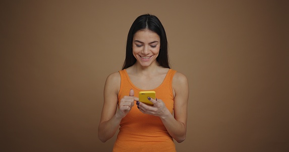 A pretty smiling woman is using a smartphone isolated on brown background