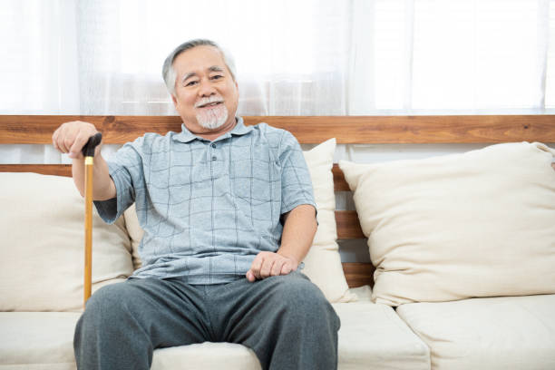 elderly old senior man sitting, resting his hands on wooden walking stick sitting on couch in living room in house after retirement. elderly old senior man sitting, resting his hands on wooden walking stick sitting on couch in living room in house after retirement. Wicker stock pictures, royalty-free photos & images