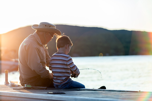 A grandfather is teaching his grandson to fish during sunset in summer. They are both sitting on the dock and are concentrated on their activity. It is a beautiful summer day. Across the lake, there is a mountain.