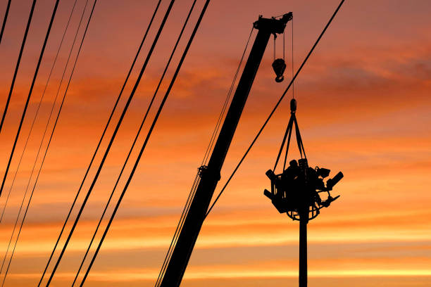 istock Silhouette mobile crane lifting electrician in telescopic boom to install electrical system on spotlights tower in sunset sky 1205879956