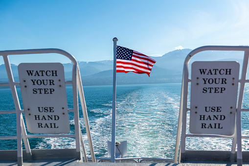 An American flag flaps in the wind at the stern of a ship, past signs warning to watch your step and use hand rail. Location: off the coast of Port Angeles, Washington State