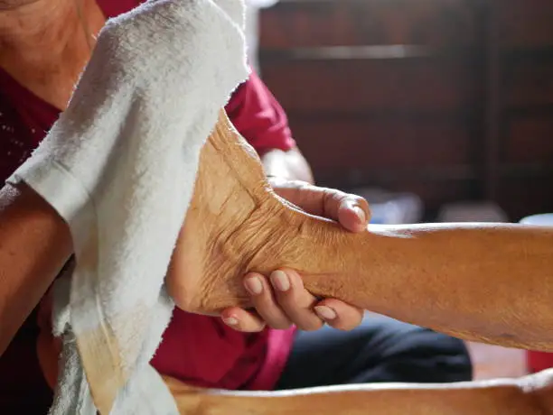 Hands of a woman holding an older person's feet, while gently wiping / cleaning it with a wet washcloth - giving an elderly a bed bath at home