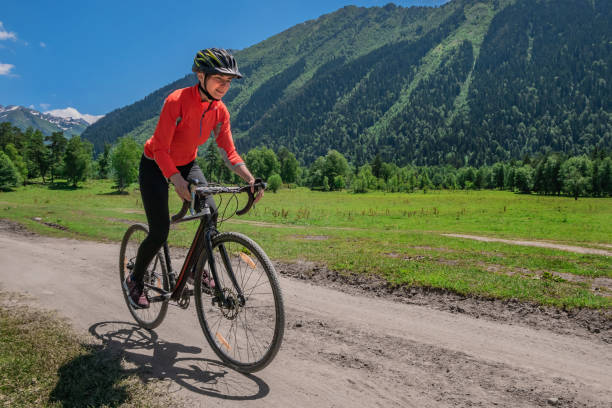 una joven sonriente en una bicicleta de ciclocross cabalga a lo largo de un sinuoso camino de montaña contra un fondo de bosque verde y montañas con glaciares y nieve en las cimas - cyclo cross fotografías e imágenes de stock