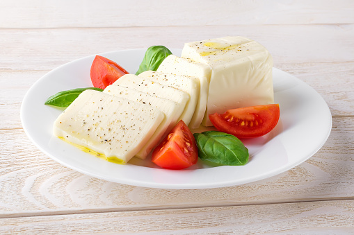Rectangular slices of mozzarella cheese, fresh basil and quarters of tomato on a white plate over a wooden table. Healhy mediterranean cuisine and vegetarian food. Front view.