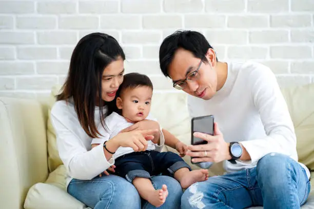 Photo of Cheerful young Asian family with son watching video on mobile phone and sitting on sofa couch at home