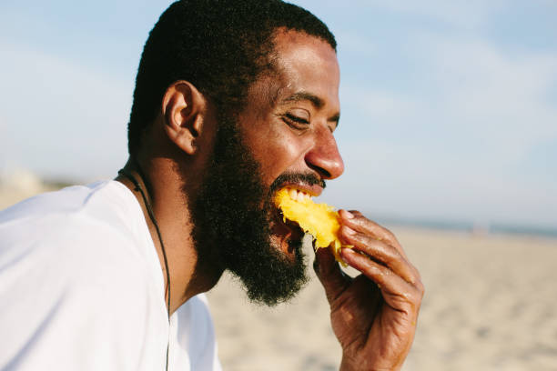 istock Man with beard and dark skin eating mango on sea shore 1205939664