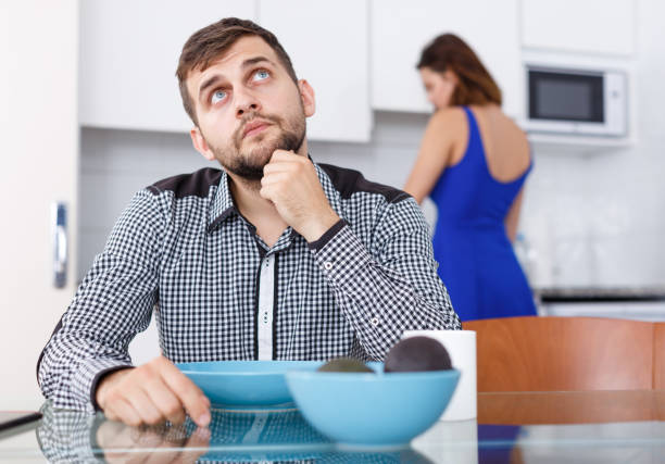 istock Young male sitting at kitchen table while woman preparing food 1205946620