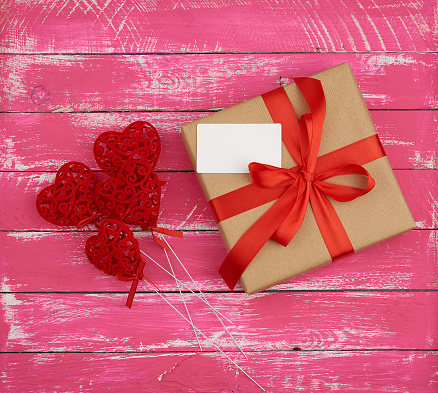 rectangular box with a gift tied with a red silk ribbon and red hearts on a pink wooden background, top view