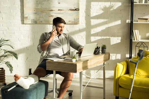 Handsome freelancer with broken leg using laptop and talking on smartphone at table in living room Handsome freelancer with broken leg using laptop and talking on smartphone at table in living room broken leg stock pictures, royalty-free photos & images