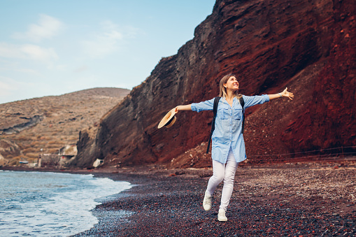 Traveler woman running along shore of Red beach on Santorini island, Greece feeling free and happy with arms opened. Traveling and vacation