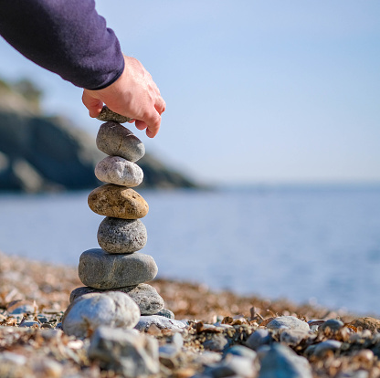 Close up of young man's hand lays a stone on a stone cairn outdoors. Pebble beach on blue sky blurry background. Winter holidays or vacation at sea in Europe. The concept of peace, tranquility.