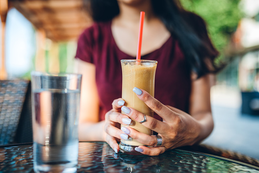 Gypsy young woman holding glass of ice coffee in cafe, close up.