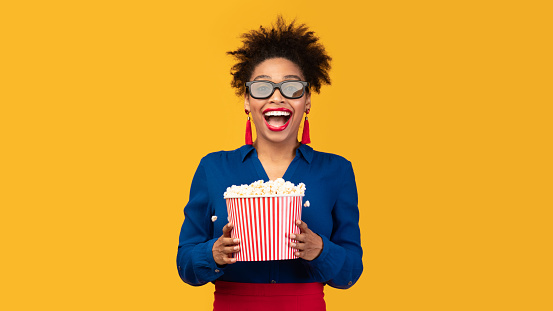 Cinema Day. Young afro woman watching interesting movie in 3d glasses, holding bucket with pop corn, copyspace