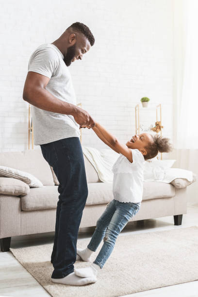 Playful daughter staying on dad feet, learning dancing Playful black daughter staying on dad feet, learning dancing at home african father stock pictures, royalty-free photos & images