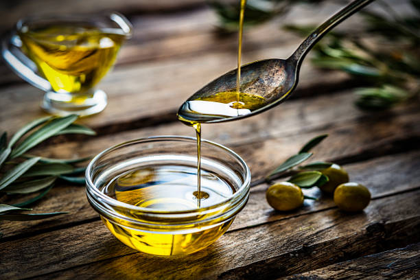 Pouring extra virgin olive oil from a vintage spoon to a glass container. Some olive branches comes from the left and right. The composition is on a rustic wooden kitchen table. Predominant colors are gold, green and brown. High resolution 42Mp studio digital capture taken with Sony A7rII and Sony FE 90mm f2.8 macro G OSS lens