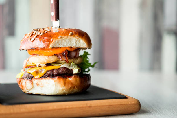 Fresh flame grilled bacon cheeseburger on wooden board Close up image of a freshly flame grilled beef burger loaded with melted cheese and crispy bacon with fresh green salad leaves and onion rings, sandwiched between a toasted sesame seed bun. The burger is displayed on a wooden board on a restaurant table with defocused background. Room for copy space. bacon cheeseburger stock pictures, royalty-free photos & images