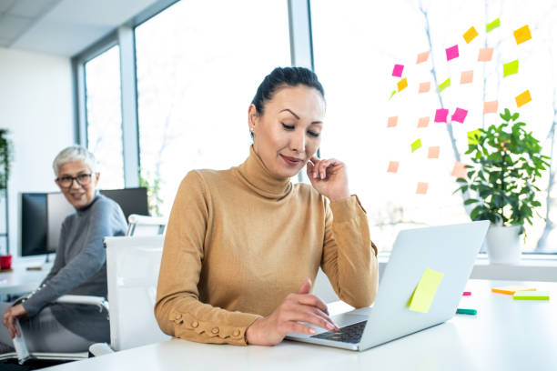 istock Two employees working in an office with a casual work model. The woman in the front is working on her lap top and the woman behind her turned to her as if she's telling her something. 1206002041