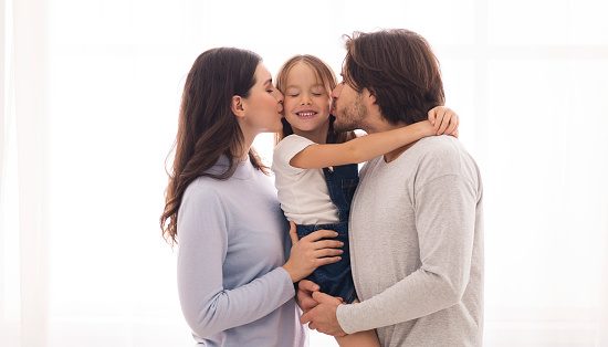 Family Care. Loving Parents Kissing Their Cute Little Daughter From Both Sides, Standing Against Window At Home, Panorama With Copy Space