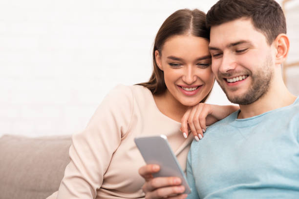 istock Smiling Couple Using Mobile Phone Sitting On Sofa Indoor 1206004742