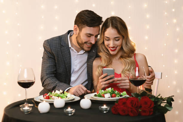 Young Couple Using Smartphone Together During Romantic Dinner In Restaurant