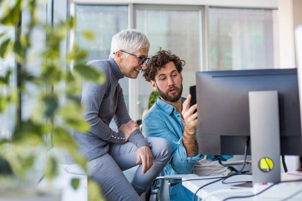 istock Two co-workers talking on a work break at the office. The man is holding a cell phone and the woman is sitting at the window sill. 1206009580