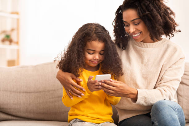 istock Afro Mother And Daughter Using Mobile Phone Sitting On Sofa 1206009620
