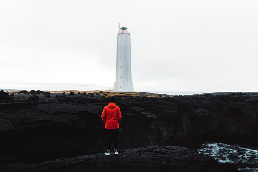 Young man tourist with red jacket walking near big white lighthouse enjoying the prefect windy day at the seashore in East Iceland