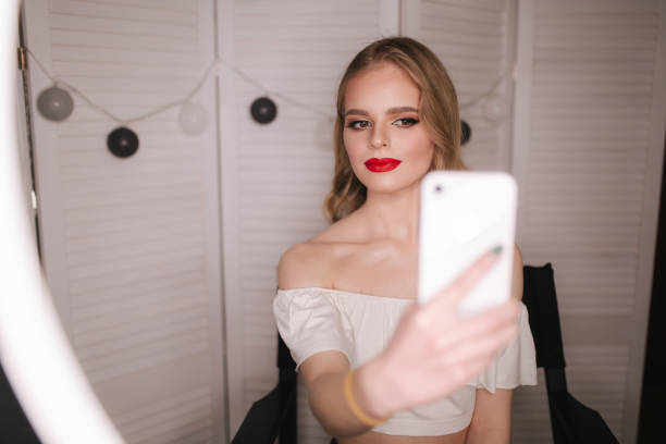 istock Young attractive model make a selfie after makeup in beauty studio. She use a ring lapm for photo. Background of white folding screen. Red lipstick 1206022738