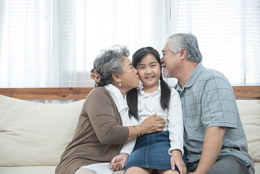 Asian grandparents kiss granddaughter cheek at home. Senior Chinese,old generation, grandfather and grandmother using family time relax with young girl kid lying on sofa in living room concept.