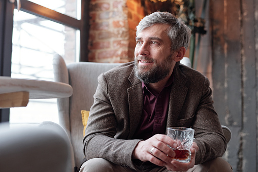 Positive dreamy middle-aged bearded man in brown jacket sitting in armchair and drinking alcohol