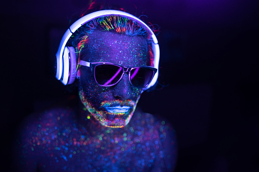 Man with glowing makeup in black light. Man with neon makeup powder on face. Man painted in fluorescent UV colors, with sunglasses and headset.