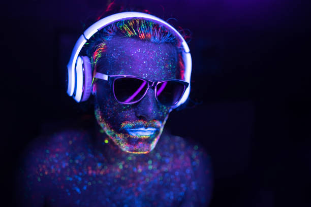 Man painted in fluorescent UV colors with sunglasses and headset Man with glowing makeup in black light. Man with neon makeup powder on face. Man painted in fluorescent UV colors, with sunglasses and headset. entertainment club photos stock pictures, royalty-free photos & images