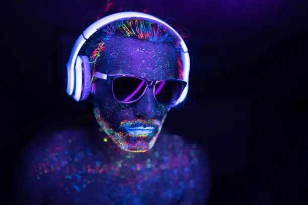 Man with glowing makeup in black light. Man with neon makeup powder on face. Man painted in fluorescent UV colors, with sunglasses and headset.
