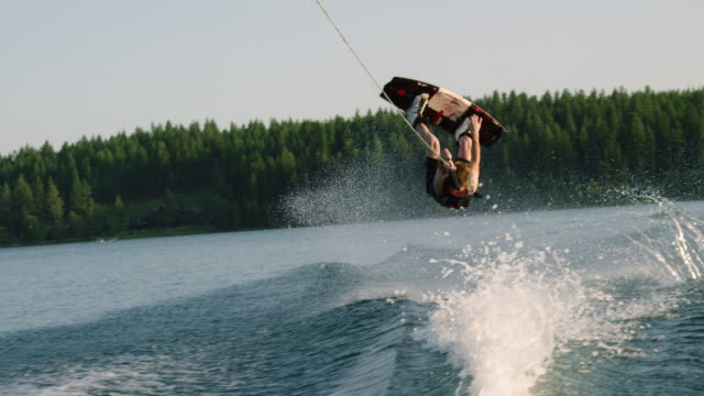 A Twenty-Something Caucasian Male Jumps and Performs Tricks on a Wakeboard before Falling on Glen Lake near Victor, Montana on a Sunny Afternoon