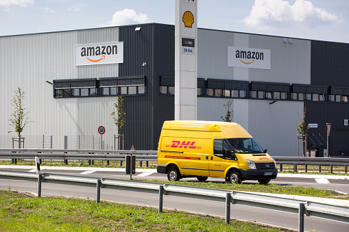 Raunheim, Germany - August 21, 2019: Facade and logo of amazon logistics center in Raunheim-Moenchhof, Germany. Amazon.com, Inc. is an American electronic commerce and cloud computing company and the largest Internet retailer in the world as measured by revenue and market capitalization.  In the foreground a passing delivery van of DHL