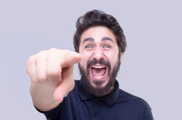 Portrait of a sneering young man looking at camera by pointing at camera over gray background Portrait of a sneering young man looking at camera by pointing at camera over gray background. Horizontal composition. Studio shot. teasing stock pictures, royalty-free photos & images