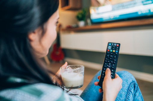 Unrecognizable young woman holding cup with hot drink and remote control