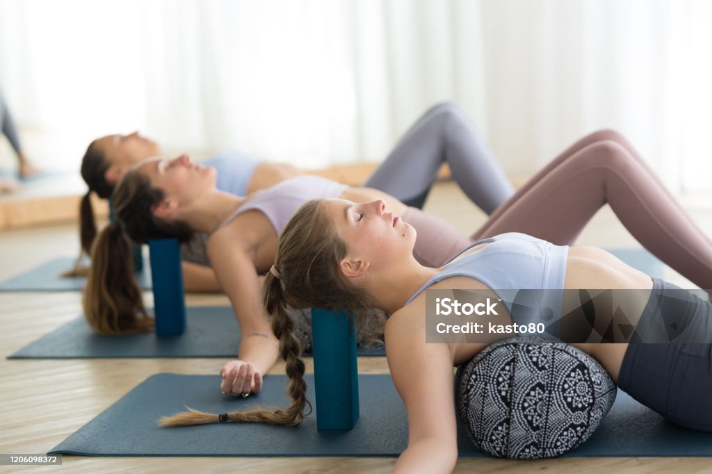 Restaurativeyoga mit einer Stütze. Gruppe von drei jungen sportlichen attraktiven Frauen im Yoga-Studio, liegend auf Bolsterkissen, Dehnen und Entspannen beim erholsamen Yoga. Gesunder aktiver Lebensstil - Lizenzfrei Yoga Stock-Foto