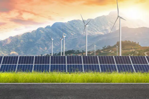 Photo of Asphalt road with solar panels with wind turbines against mountanis landscape against sunset sky,Alternative energy concept