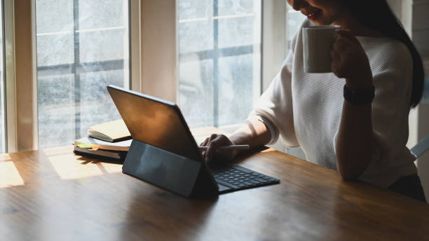 istock Closeup woman holding a coffee cup in hand while using a computer tablet and sitting at the modern working desk in the morning with comfortable living room and windows as background. 1206150253