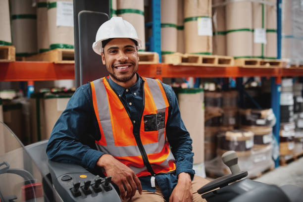 istock Handsome engineer wearing protective uniform and hardhat smiling joyfully to the camera sitting in forklift stacker while working at the storage 1206156645