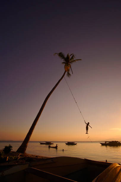 silhouette von indentify bajau kids spielen eine schaukel unter kokosnussbaum bei sonnenuntergang auf maiga island, sempoerna - indentify stock-fotos und bilder