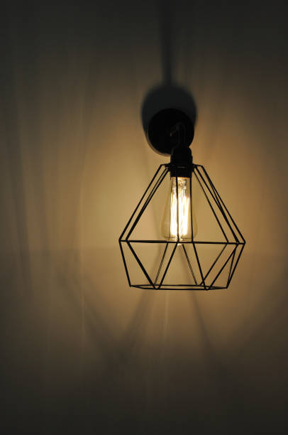 istock Modern pendant light closeup. Diamond shape wire lampshade with light bulb inside. Glowing filament in lamp of minimal style. Sconce lamp hanging on wall background with copy space. Interior lighting. 1206182371