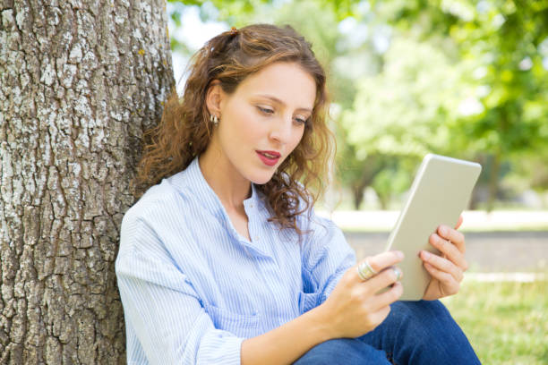 istock Thoughtful young woman reading from tablet in park 1206184520
