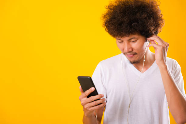 Happy asian handsome man with curly hair he smiling enjoying listening to music on earphones using a mobile smartphone Happy asian handsome man with curly hair he smiling enjoying listening to music on earphones using a mobile smartphone isolate on yellow background asian man headphones stock pictures, royalty-free photos & images